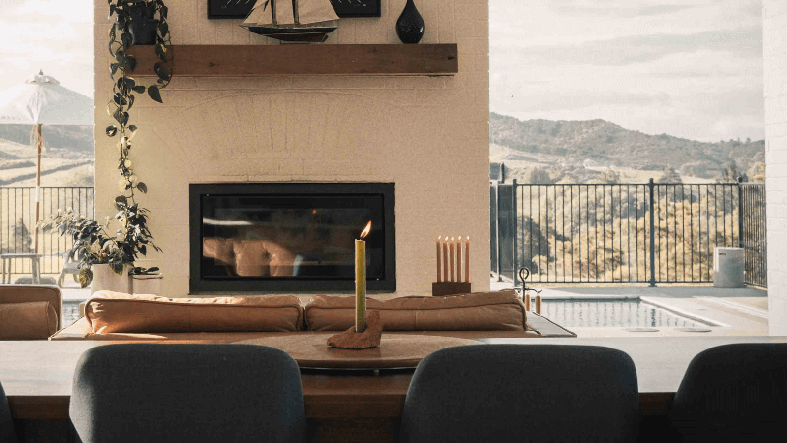 An open plan living space, with a table and chairs in the foreground, with a fireplace set between two floor to ceiling windows in the background. There is a view to hills out the windows. A recycled red brick Shard Candle Holder sits on the table and houses an olive candle stick. A recycled red brick Centrepiece Candle Holder is in the background sitting on the fire hearth. This candle holder houses 5 Terracotta candlesticks.