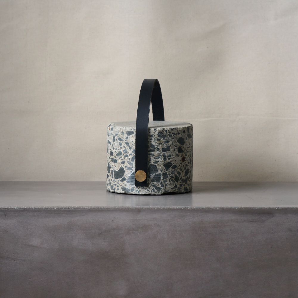 Concrete cylindrical doorstop. A black strap arches over the top and acts as a handle. The strap is bolted in at either side with brass bolts.