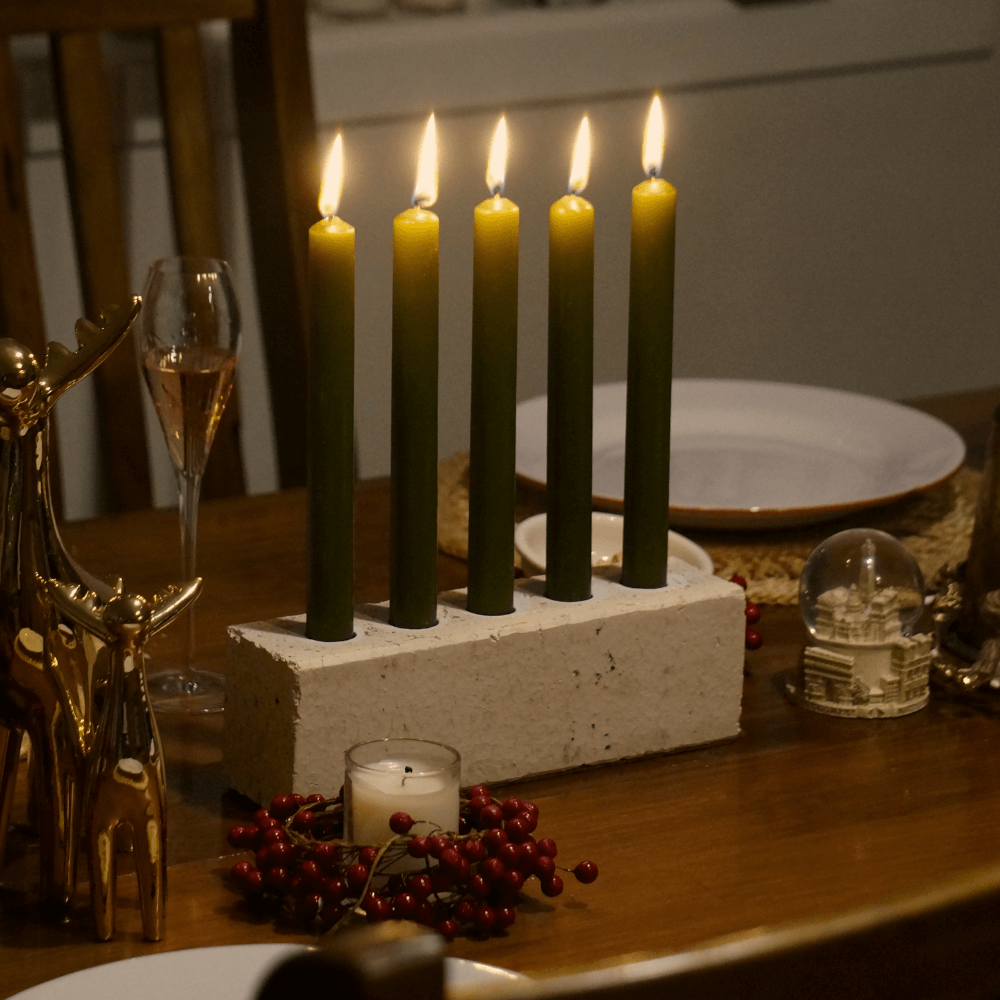 White centrepiece candleholder on a dining table. The candleholder is made from a recycled sand brick. It houses 5 candles in the olive colour way. The candles have been lit.