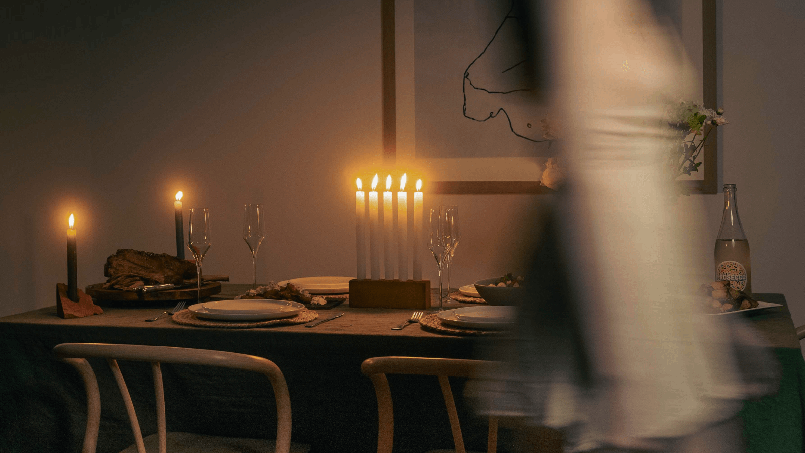 A dimly lit dinner party scene featuring a table setting for 4 people, including placemats, plates, wine glasses and cutlery. On the middle of the table sits The Centrepiece recycled brick candle holder which houses 5 lit white candle sticks. 2 lit recycled brick The Shard Candle Holders are also on the table. A woman has walked across the foreground of the photo and is blurry due to the photographs long exposure time. 